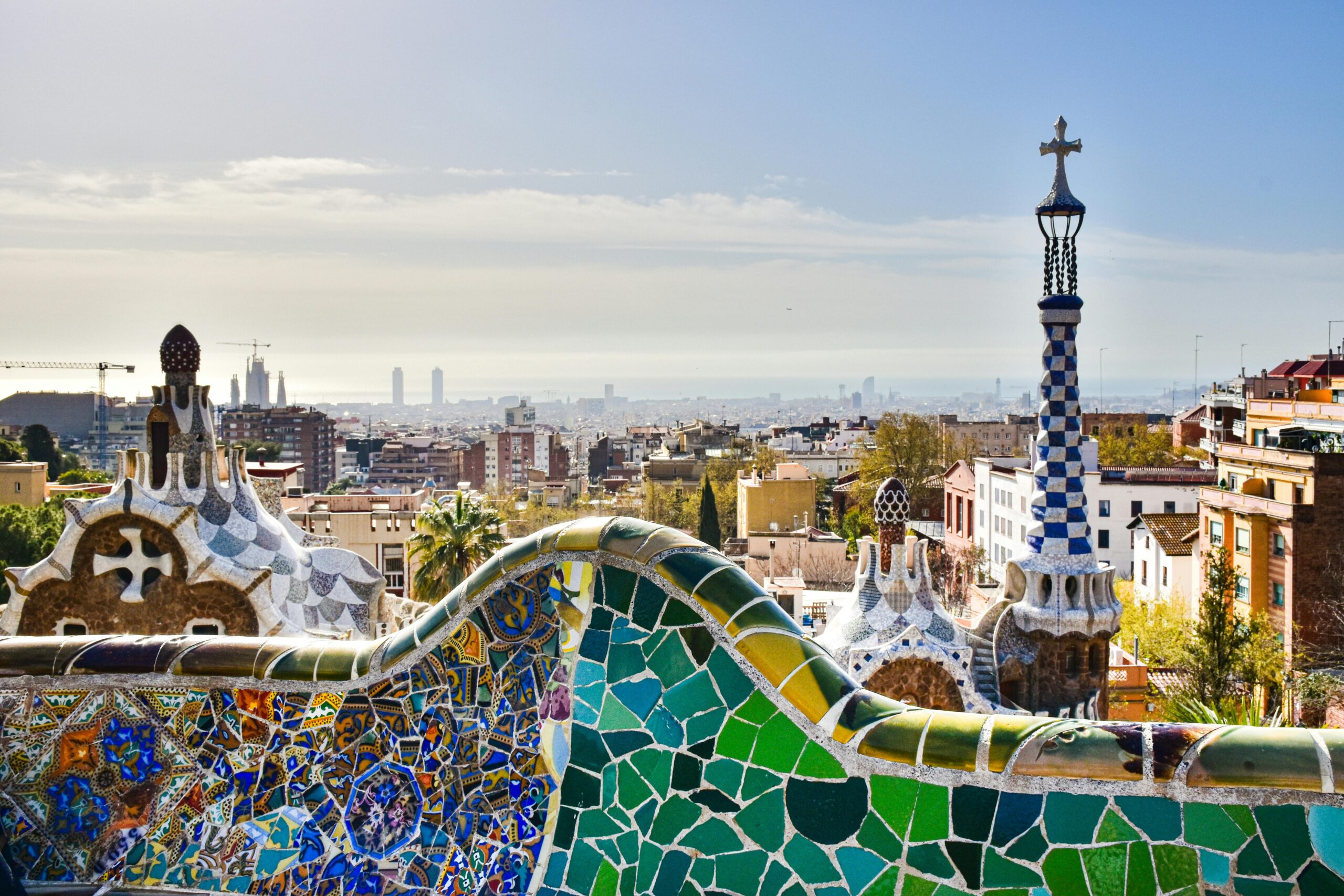 Colorful mosaic bench and panoramic view of Barcelona from Park Güell, with Gaudí's architecture in the foreground.